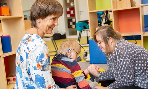Eine Szene vom integrativen Kindergarten und Hort in Vöcklabruck. Eine Frau und zwei Kinder mit Beeinträchtigung sitzen am Boden in einem Spielzimmer