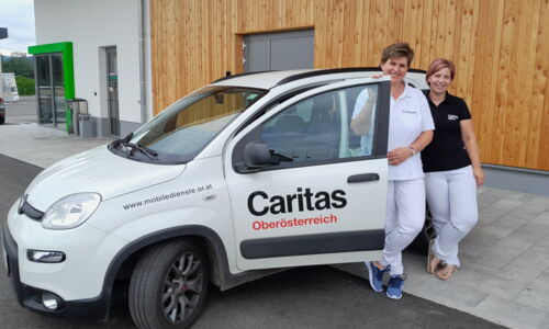 Zwei Pflegekräfte von der Caritas stehen vor einem PKW der Caritas OÖ