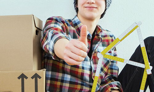 Ein Jugendlicher sitzt neben Umzugskartons und hält einen Zollstock, der wie ein Haus geformt ist.
