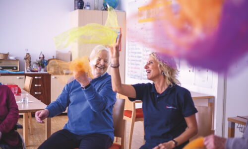 In einem Gruppenraum sitzen ein älterer Herr und und eine Frau im Sesselkreis, sie werfen bunte Tücher in die Luft