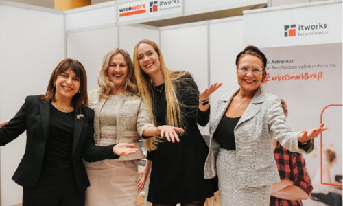 Frauen stehen bei einem Messestand und blicken mit einladender Geste in die Kamera