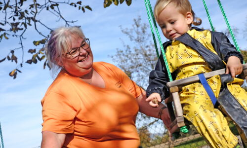 Eine Frau im orangen Caritas T-Shirt hilft einem Kleinkind beim Schaukeln
