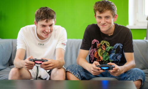 Zwei Burschen sitzen auf einer Couch und halten die Steuerung für eine Spielkonsole