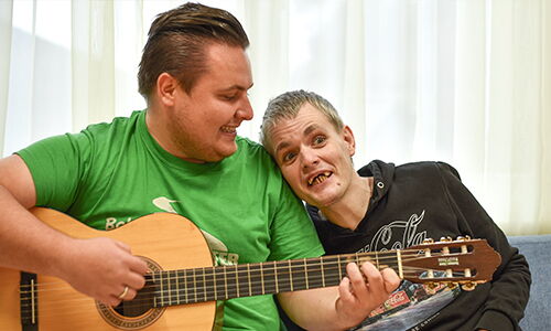 Ein Mann spielt Gitarre, ein anderer Mann lehnt sich an seine Schulter und singt.