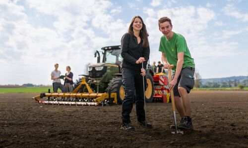 Ein Mädchen und ein Junge stehen auf einem Feld, im Hintergrund ist ein Traktor zu sehen.