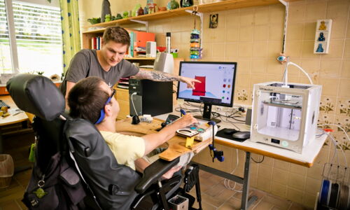 Ein junger Mann arbeitet am Computer, er sitzt im Rollstuhl. Ein anderer junger Mann zeigt auf den Bildschirm.