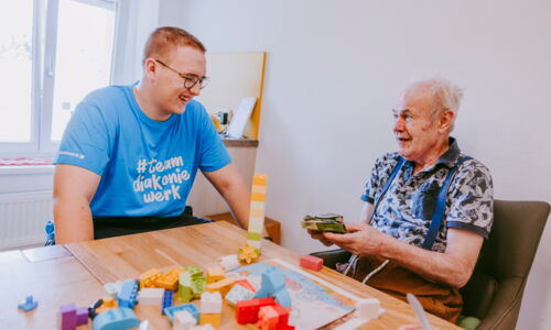 Ein junger Betreuer vom Diakoniewerk im blauen T-Shirt spielt mit einem älteren Herren verschiedene Spiele